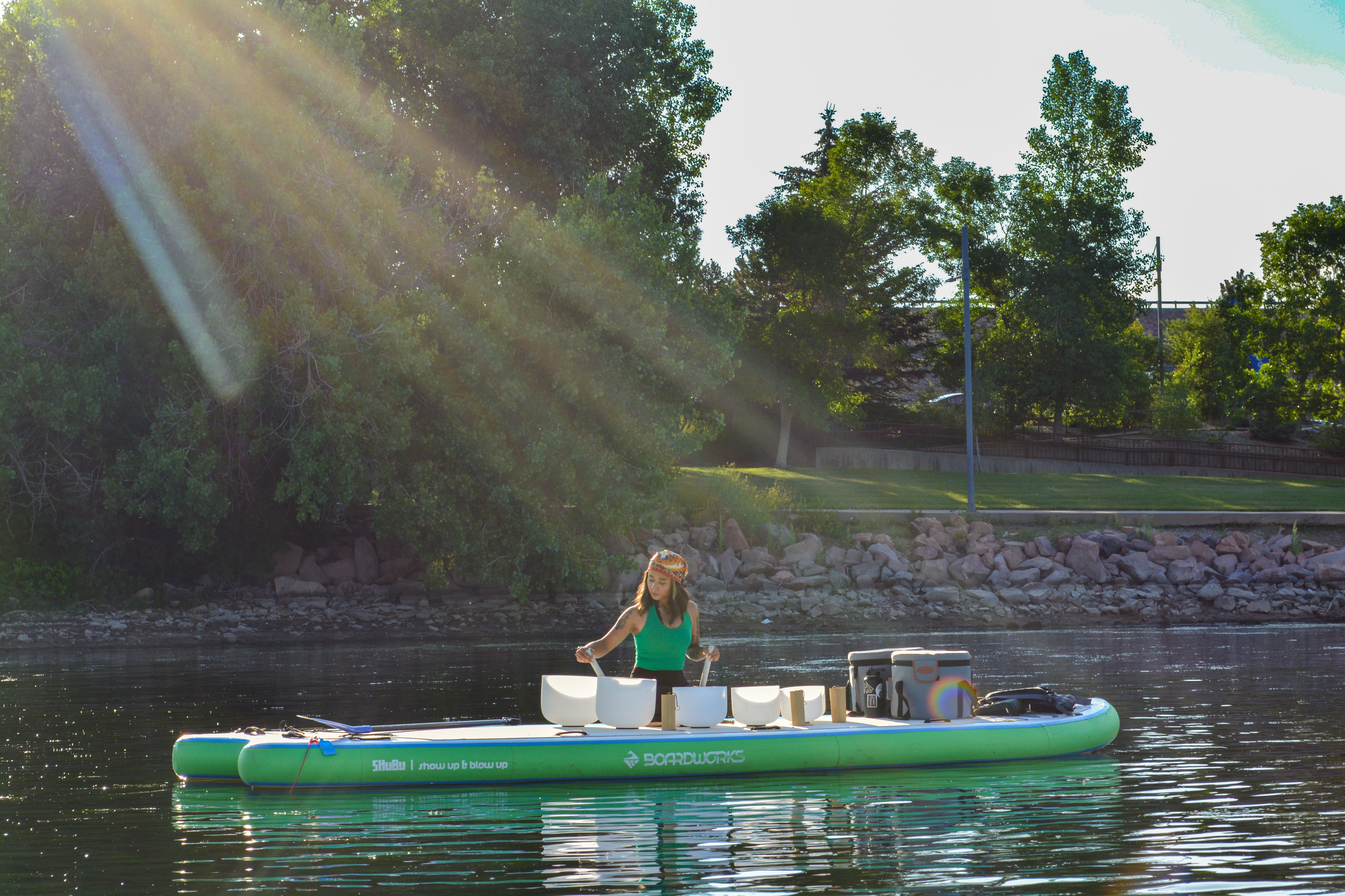 Person playing crystal singing bowls in a floating sound bath on a green inflatable paddleboard at a sunlit lake with a tree-lined park shoreline