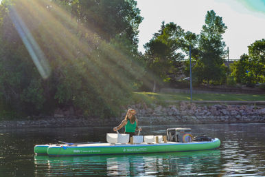 Person playing crystal singing bowls in a floating sound bath on a green inflatable paddleboard at a sunlit lake with a tree-lined park shoreline