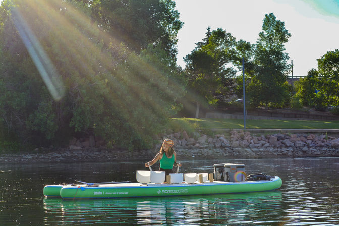 Person playing crystal singing bowls in a floating sound bath on a green inflatable paddleboard at a sunlit lake with a tree-lined park shoreline