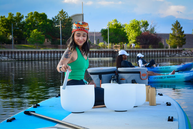 Person in a colorful headband tapping white crystal singing bowls on a paddleboard for a lakeside sound bath, calm water with kayakers and a tree-lined waterfront in the background.
