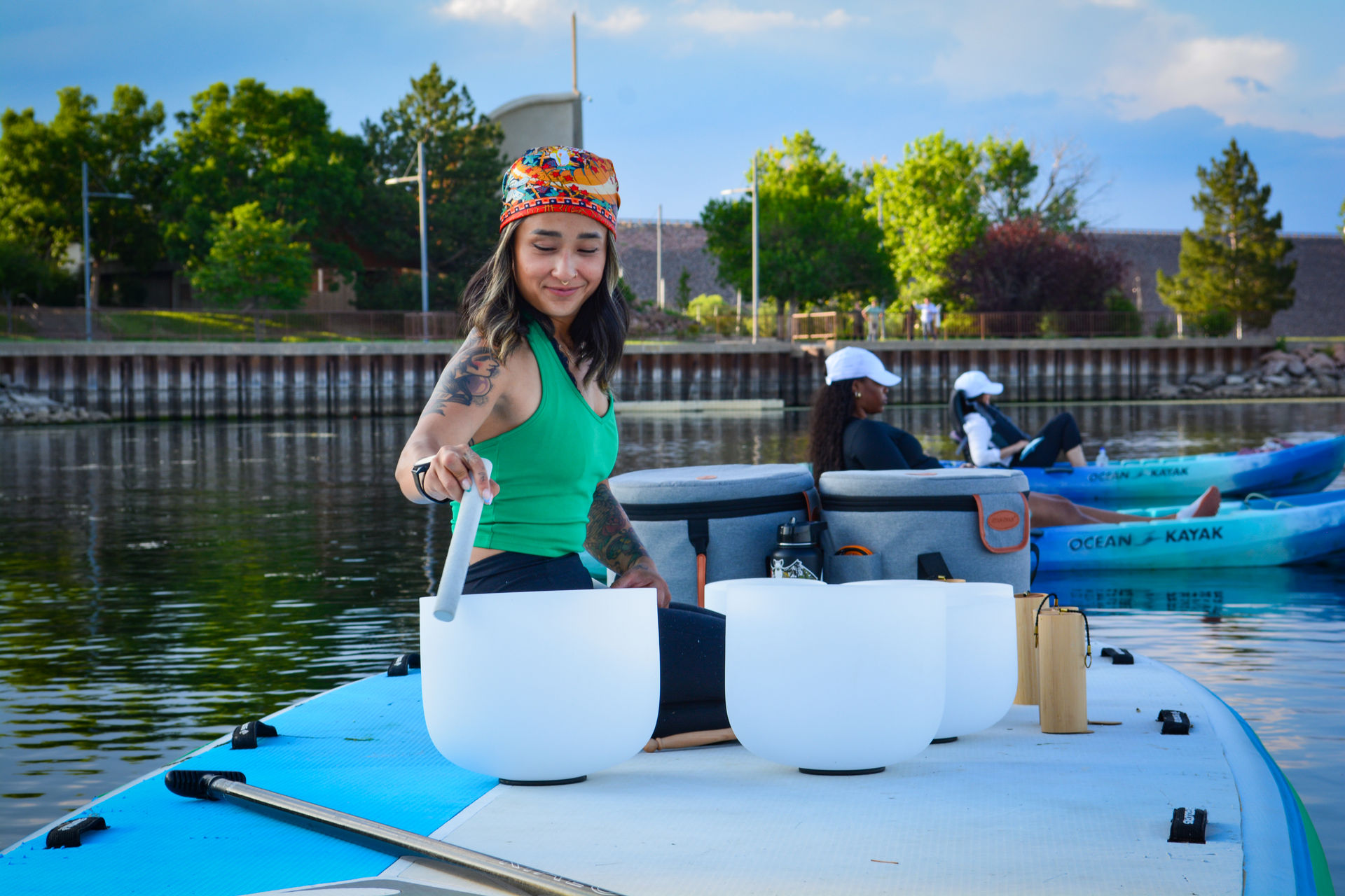 Person in a colorful headband tapping white crystal singing bowls on a paddleboard for a lakeside sound bath, calm water with kayakers and a tree-lined waterfront in the background.