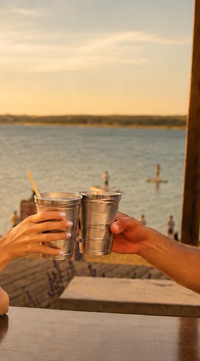 Two friends in sunglasses clinking metal cups on a lakeside patio at sunset, paddleboarders and a sandy shore visible in the background