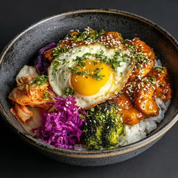 Korean-style rice bowl with white rice, saucy spicy chicken, sunny-side-up egg, kimchi, pickled purple cabbage and charred broccoli, topped with sesame seeds and chopped herbs