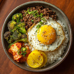 Korean-style rice bowl with two sunny-side-up eggs sprinkled with sesame seeds and scallions, seasoned ground beef, roasted broccoli, spicy kimchi and pickled radish over steamed white rice in a ceramic bowl