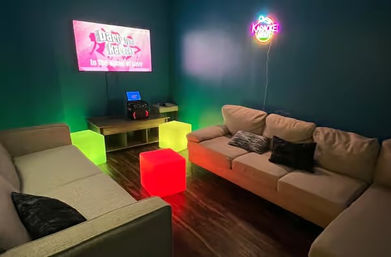 Cozy private karaoke room with two beige sofas facing a wall-mounted TV showing a karaoke screen, neon karaoke sign on the wall, glowing red and green LED cube seats and dark hardwood floor.