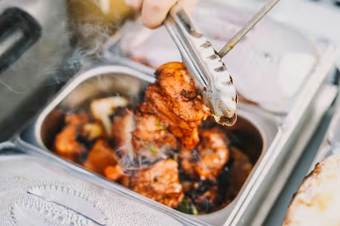 Steaming, sizzling spiced grilled chicken pieces lifted with tongs from a hot stainless-steel serving tray at a bustling street-food or buffet counter.