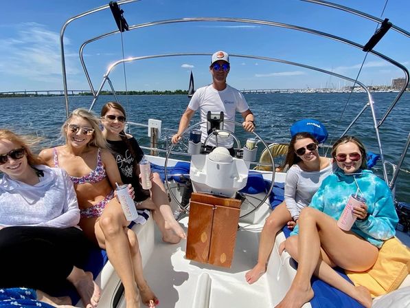 Six friends smiling and holding tumblers while lounging on a sailboat, skipper at the wheel, sunny blue sky, calm harbor and bridge in the background.
