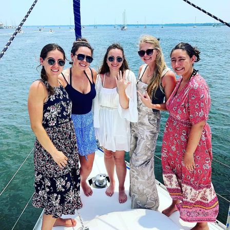 Five women in summer dresses and sunglasses smiling on a sailboat deck, one showing a ring, with sailboats and a coastal harbor in the background.