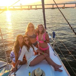 Four women in swimsuits smiling on a sailboat bow at golden sunset, calm harbor waters and a bridge in the background.