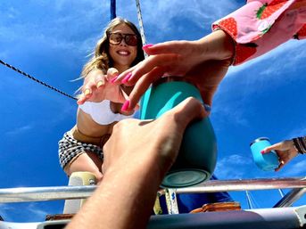 Smiling woman on a sailboat deck reaches down to hand a turquoise insulated cup to someone below, bright blue sky and colorful manicured nails visible.
