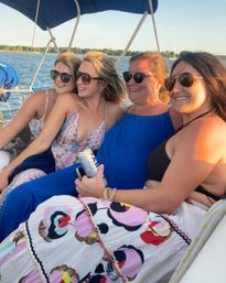 Four friends in sunglasses smiling and lounging on a sailboat over a calm lake at sunset, wearing summer dresses and holding a canned drink.