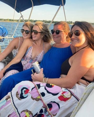 Four friends in sunglasses smiling and lounging on a sailboat over a calm lake at sunset, wearing summer dresses and holding a canned drink.