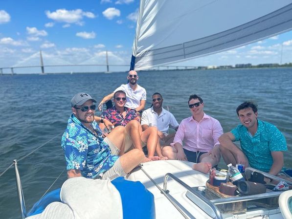 Six friends relaxing on a sailboat under bright blue skies, wearing colorful shirts and sunglasses, sailing on calm coastal waters with a long bridge on the horizon.
