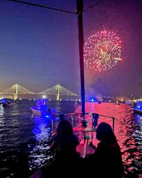Fireworks burst over an illuminated cable-stayed bridge at dusk as a crowd of boats gathers in the harbor, red and blue lights reflecting on the water and two silhouetted people watching from a boat bow.