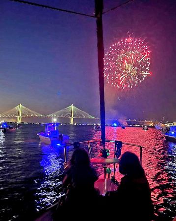 Fireworks burst over an illuminated cable-stayed bridge at dusk as a crowd of boats gathers in the harbor, red and blue lights reflecting on the water and two silhouetted people watching from a boat bow.