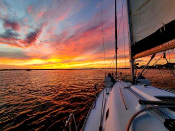 Sailboat bow on a sunset sail with vivid orange-pink clouds reflecting on rippling coastal water, birds flying overhead and a distant shoreline on the horizon.