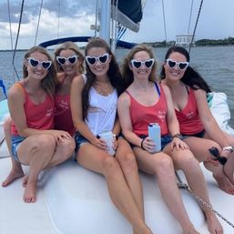 Five women in matching tank tops and white heart-shaped sunglasses smiling on a sailboat in a coastal bay, holding drinks — bachelorette/girls' trip