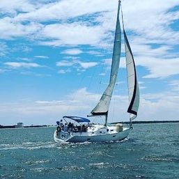 White sailboat with tall gray-and-white sails and a blue cockpit canopy gliding on turquoise coastal waters near a distant shoreline under a bright partly‑cloudy sky