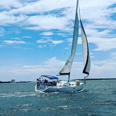White sailboat with tall gray-and-white sails and a blue cockpit canopy gliding on turquoise coastal waters near a distant shoreline under a bright partly‑cloudy sky