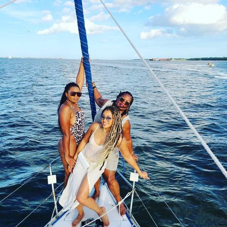 Three friends posing on the bow of a sailboat during a sunny summer sail in blue coastal waters, smiling and holding the mast with a distant shoreline under a partly cloudy sky.