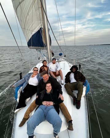 Six friends lounging on the deck of a white sailboat with its mainsail up, cruising calm open water under a pastel cloudy sky.