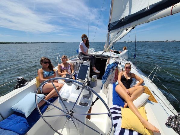 Four friends relaxing on a sunlit sailboat deck, lounging on cushions and towels near the helm with white sails billowing over calm coastal waters and a distant shoreline.