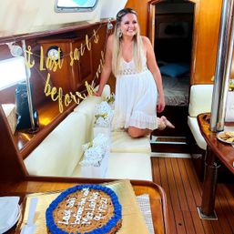 Smiling woman in a white sundress kneeling on a cushioned bench inside a wooden sailboat cabin, surrounded by a gold celebration banner, gift bags, and a large cookie cake with blue icing on the table — onboard yacht party.
