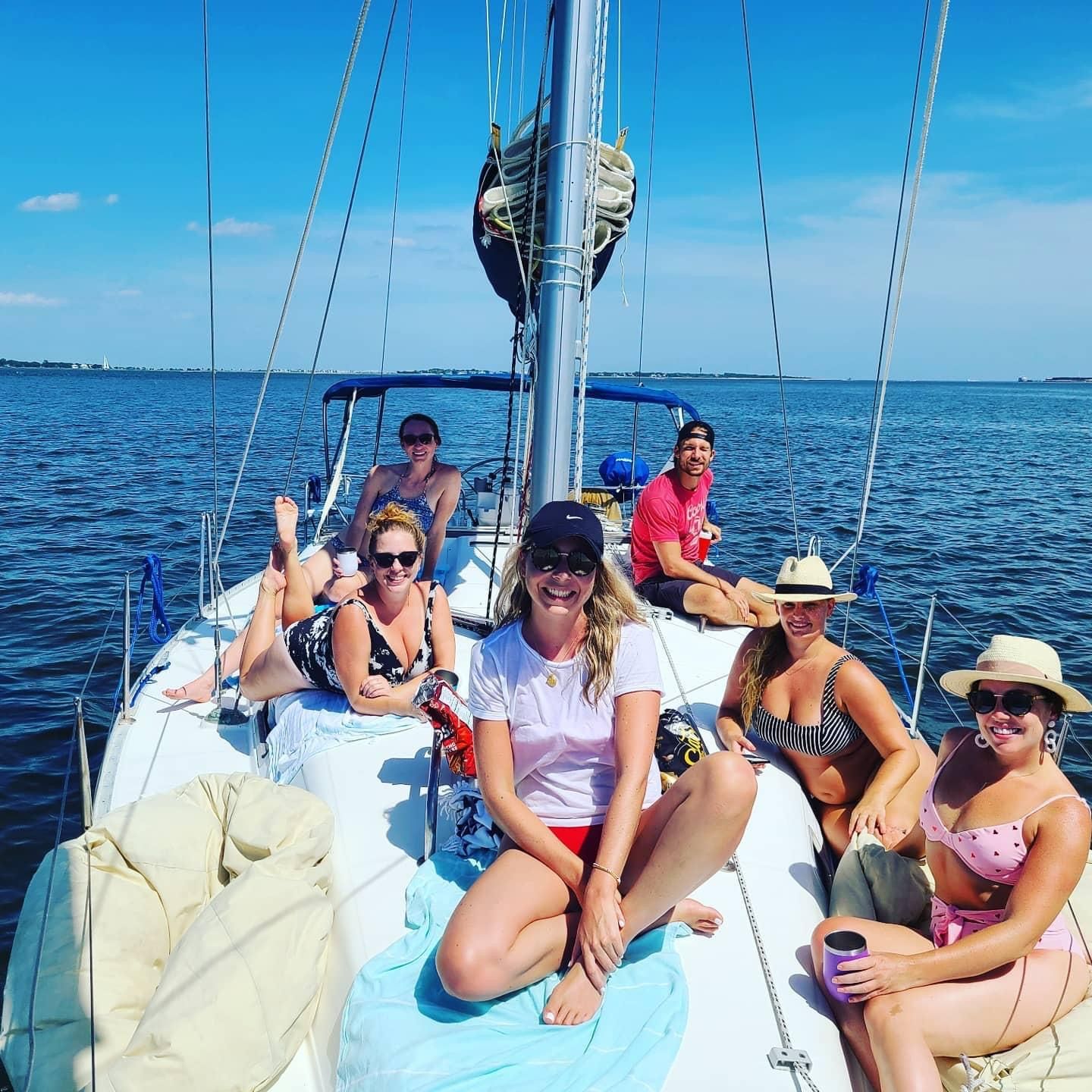 Group of six friends smiling and relaxing on the deck of a sailboat in calm coastal waters under a bright blue sky — summer sailing day with swimsuits, hats, and sunlit ocean views.