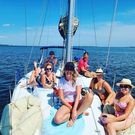 Group of six friends smiling and relaxing on the deck of a sailboat in calm coastal waters under a bright blue sky — summer sailing day with swimsuits, hats, and sunlit ocean views.