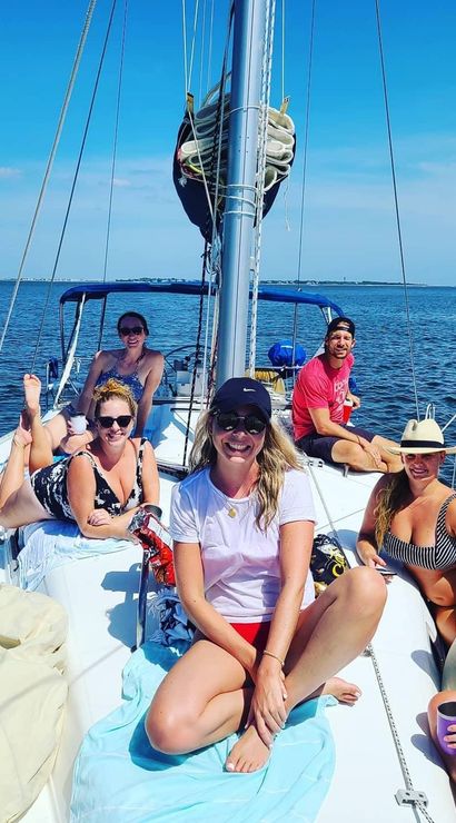Group of six friends smiling and relaxing on the deck of a sailboat in calm coastal waters under a bright blue sky — summer sailing day with swimsuits, hats, and sunlit ocean views.