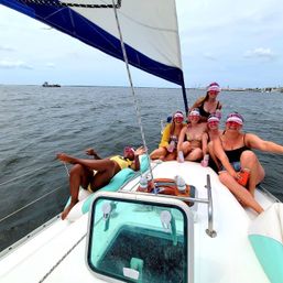 Six friends wearing pink visors laughing and holding canned drinks on the bow of a sailboat with a blue-and-white sail and distant coastal skyline