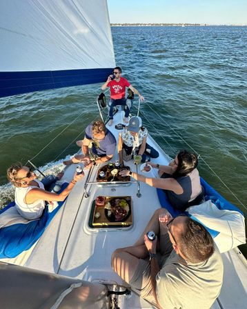 Six people relaxing on a sunlit sailboat under a white-and-blue sail, sharing drinks and a charcuterie board while sailing on coastal waters with a distant shoreline.