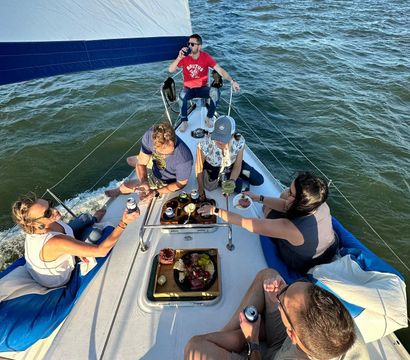 Six people relaxing on a sunlit sailboat under a white-and-blue sail, sharing drinks and a charcuterie board while sailing on coastal waters with a distant shoreline.