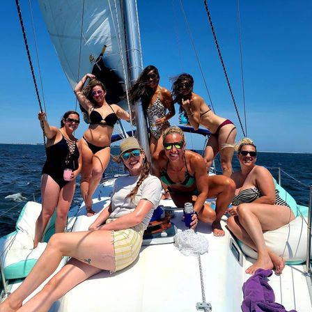 Seven friends in swimsuits smiling and holding drinks on the deck of a sailboat under full sail on a sunny summer day over blue coastal waters