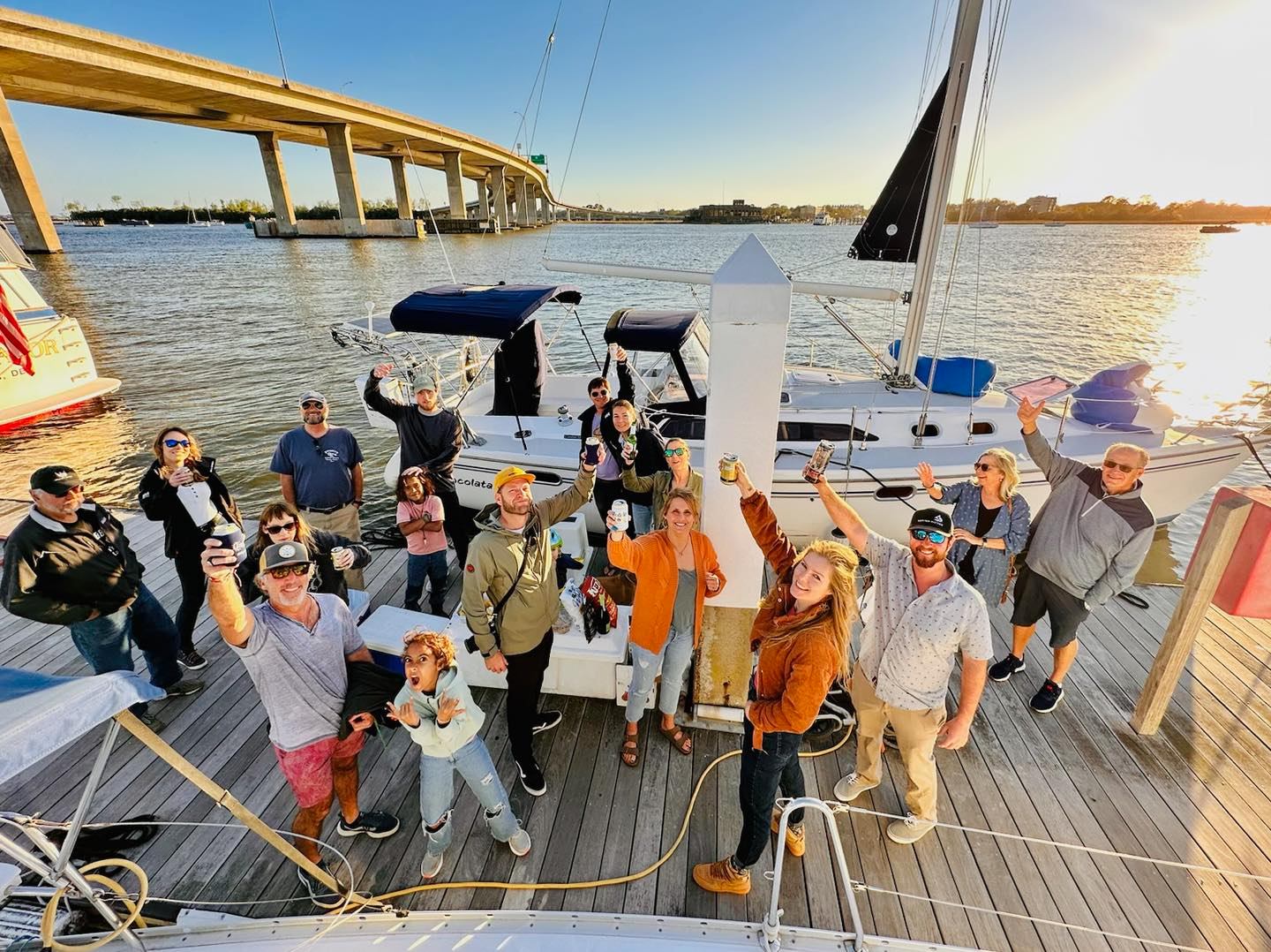 Group of friends cheering with drinks on a wooden dock beside a moored sailboat at a waterfront marina under a curved bridge at sunset