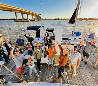 Group of friends cheering with drinks on a wooden dock beside a moored sailboat at a waterfront marina under a curved bridge at sunset
