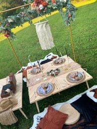 Boho outdoor picnic on a green lawn featuring low wooden tables, floral arch with macramé hanging, woven place settings with gold cutlery, black taper candles, cowhide blankets, rust pillows and a record player.