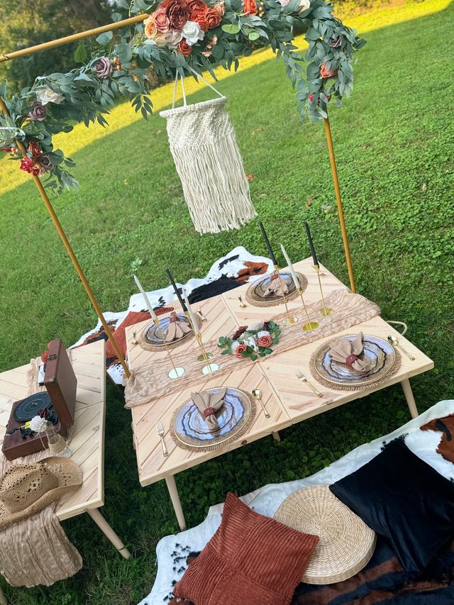 Boho outdoor picnic on a green lawn featuring low wooden tables, floral arch with macramé hanging, woven place settings with gold cutlery, black taper candles, cowhide blankets, rust pillows and a record player.