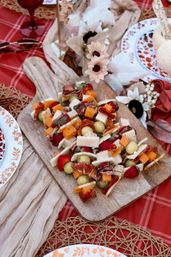 Outdoor picnic spread: rustic wooden board piled with colorful charcuterie skewers — cheddar and white cheese cubes, salami, grapes and strawberries — styled on a red plaid blanket with floral accents