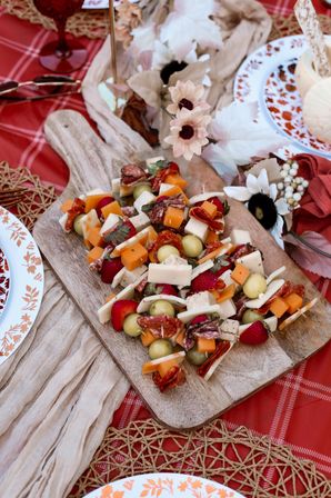 Outdoor picnic spread: rustic wooden board piled with colorful charcuterie skewers — cheddar and white cheese cubes, salami, grapes and strawberries — styled on a red plaid blanket with floral accents