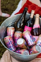 Galvanized metal ice bucket filled with pink-grapefruit sparkling water cans and assorted rosé and red wine bottles with pink labels, set on grass for an outdoor picnic.