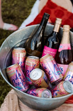 Galvanized metal ice bucket filled with pink-grapefruit sparkling water cans and assorted rosé and red wine bottles with pink labels, set on grass for an outdoor picnic.