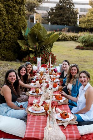 Group of friends enjoying an outdoor fall picnic in a grassy garden with a long red-plaid tablescape styled with small pumpkins, candles, amber glassware and autumn place settings.