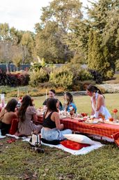 Friends enjoying an outdoor autumn picnic in a park with a low red-check table, fall tablescape, candles, pumpkins and cozy pillows