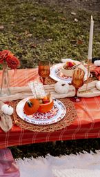 Cozy outdoor fall picnic tablescape on a red plaid tablecloth with mini pumpkin bowls of snacks, amber goblets, woven placemats, white taper candle, small decorative pumpkins and autumn florals on grass
