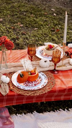Cozy outdoor fall picnic tablescape on a red plaid tablecloth with mini pumpkin bowls of snacks, amber goblets, woven placemats, white taper candle, small decorative pumpkins and autumn florals on grass