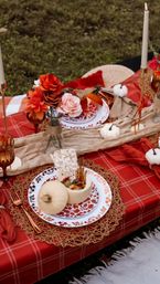Outdoor autumn picnic tablescape on grass with a red plaid tablecloth, woven placemat, patterned plates, small white pumpkins and a hollowed pumpkin bowl of snacks, crackers, copper flatware, amber goblets, tall candles and an orange-pink floral centerpiece — cozy fall picnic setting.