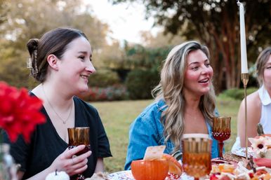 Two friends chatting and smiling at an outdoor autumn picnic table with pumpkins, amber goblets and a tall taper candle in a leafy backyard setting