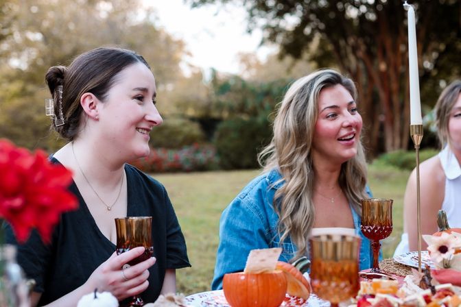 Two friends chatting and smiling at an outdoor autumn picnic table with pumpkins, amber goblets and a tall taper candle in a leafy backyard setting