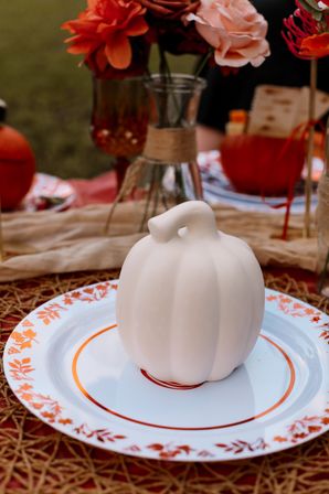 White ceramic pumpkin placed on a white plate with orange floral rim, styled as a fall tablescape centerpiece with orange flowers and rustic outdoor autumn decor.